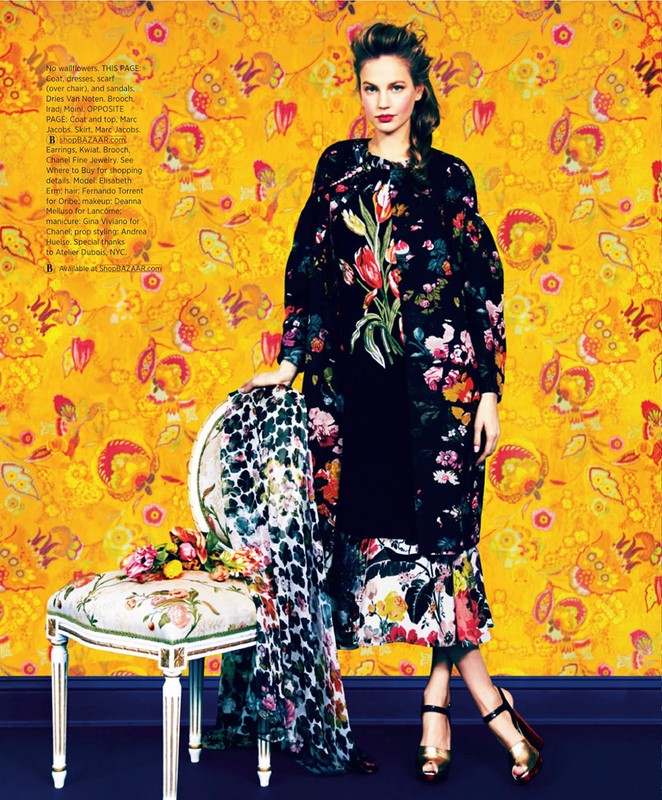Fashion editorial shot of a woman in a floral coat and gold high heels posing next to a decorated chair against a floral wallpaper backdrop.