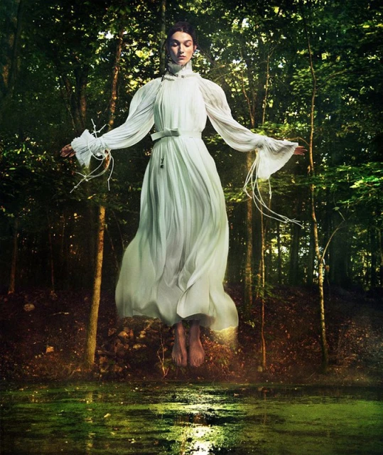 Woman in long flowing white dress levitating in a forest with pond, lit by golden light. Ethereal fashion photo.