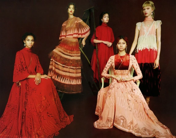 Five diverse fashion models pose in a photo studio wearing long, elegant gowns with intricate details and unique designs against a dark background.