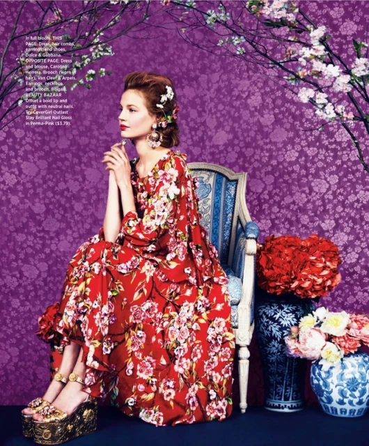 A woman in a floral dress sits on an ornate chair, surrounded by flowers and a patterned purple background, in a fashion editorial style.