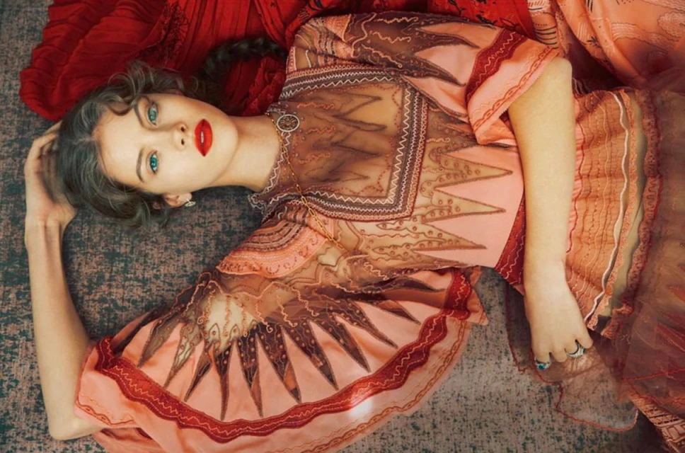 Brunette woman with red lipstick lies on a patterned surface, wearing a patterned orange top with jewelry in a high-angle shot.