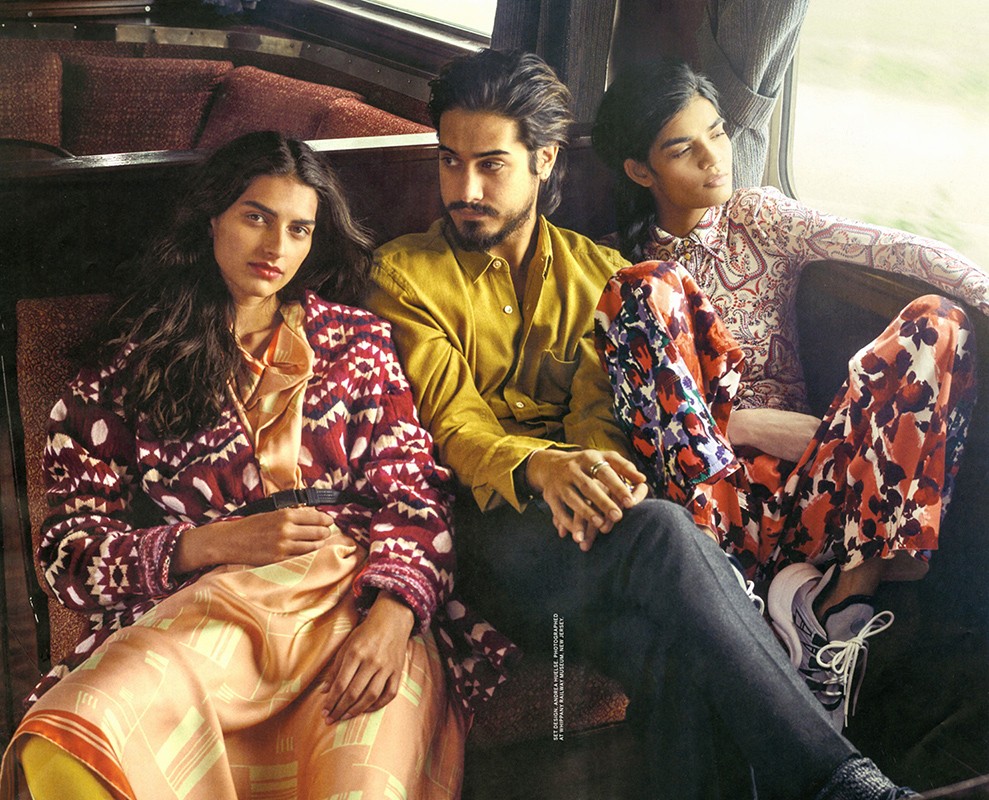 Three models seated together on a vintage train carriage bench wearing patterned clothing and a mustard shirt, styled by Andrea Huelse.