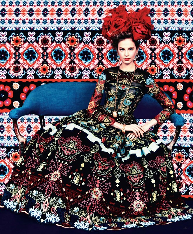 A woman in an ornate patterned dress sits on a blue couch with a colorful patterned background. Her headpiece is composed of large red flowers.