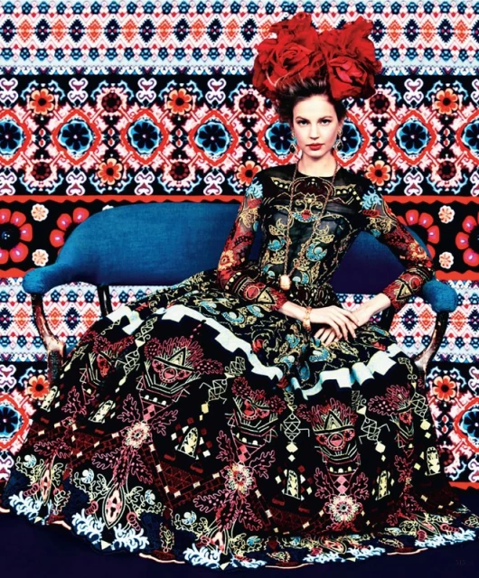 A woman in an ornate patterned dress sits on a blue couch with a colorful patterned background. Her headpiece is composed of large red flowers.