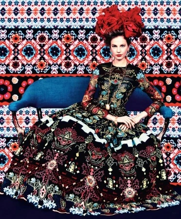 A woman in an ornate patterned dress sits on a blue couch with a colorful patterned background. Her headpiece is composed of large red flowers.