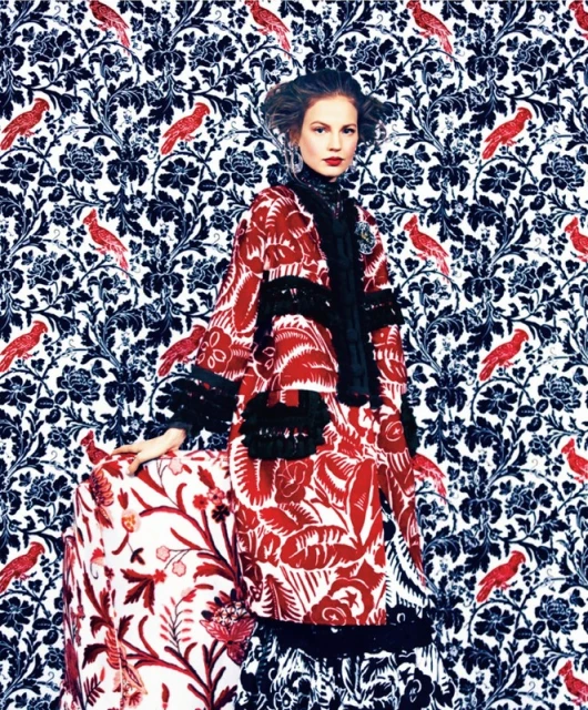 A woman in a red patterned coat stands in front of a blue floral and bird pattern, wearing an ornate hairstyle and red lipstick.