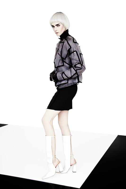 Fashion shot of an adult female with platinum blonde hair wearing a grey geometric jacket, black dress, white tights, and white boots in a studio.