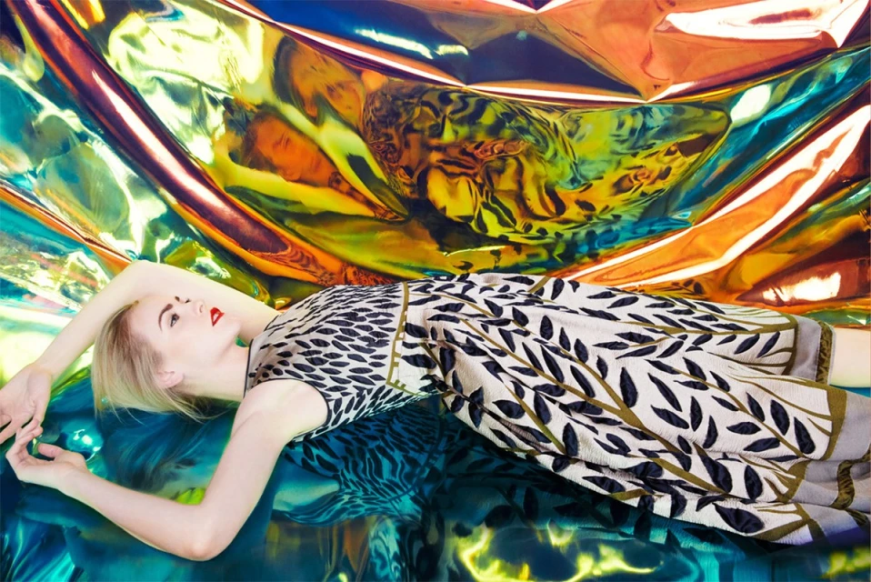 A fair-skinned woman with blonde hair lies on iridescent material in a patterned dress, with a colorful, crinkled backdrop.