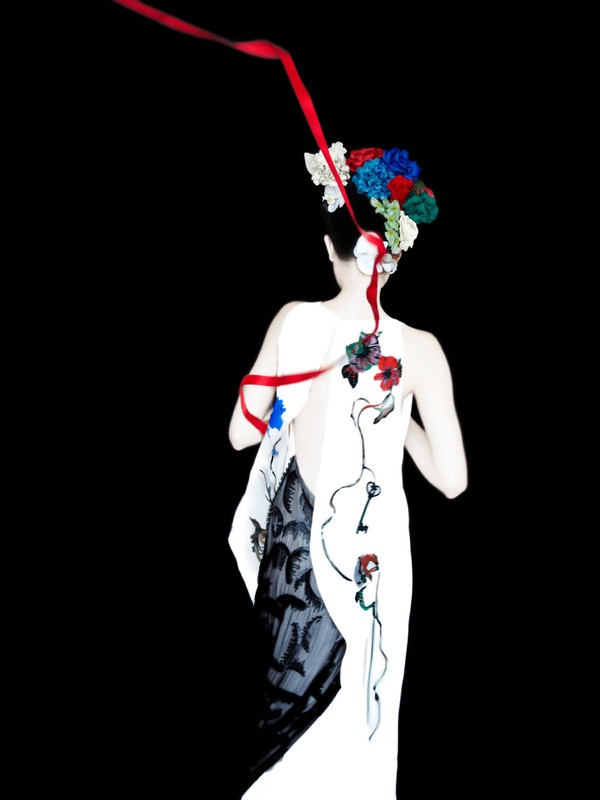 Back view of a woman in a white dress with floral accents and flowing red ribbon against a dark background in a studio setting.