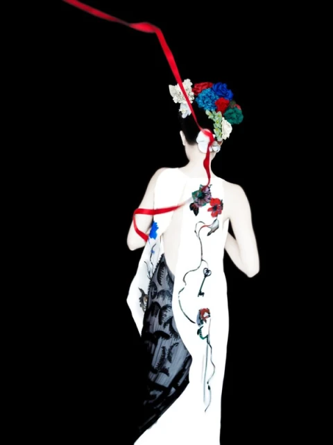 Back view of a woman in a white dress with floral accents and flowing red ribbon against a dark background in a studio setting.