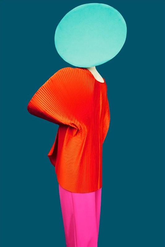 Abstract fashion image: An adult poses in a bright orange pleated top and magenta pants, with a teal disc covering their head.
