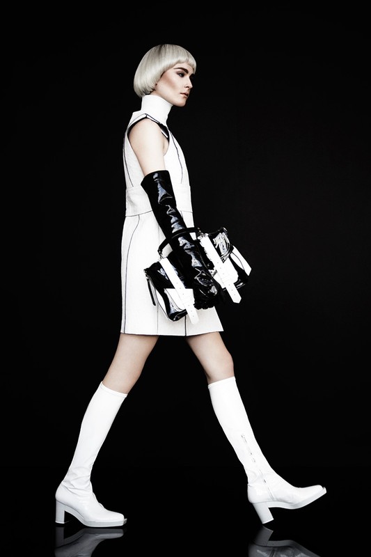 Fashion shot of a woman with a blonde bob hairstyle, wearing a white dress and boots, holding a black and white purse, against a black backdrop.