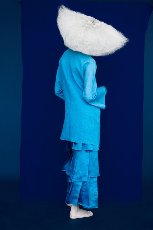 Person in blue outfit with exaggerated white headpiece stands in front of a blue background, full shot, studio lighting.