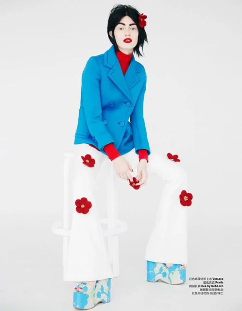 A woman in a blue blazer and white pants with red floral details sits on a white stool against a stark white backdrop in a studio shot.