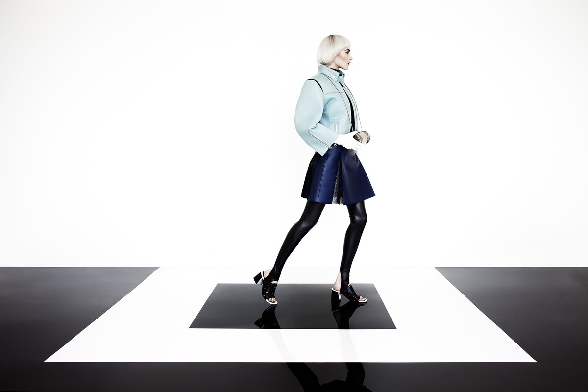 Fashion model poses in a short skirt, jacket, and heels in a studio with a black and white floor. She has white-blonde hair.