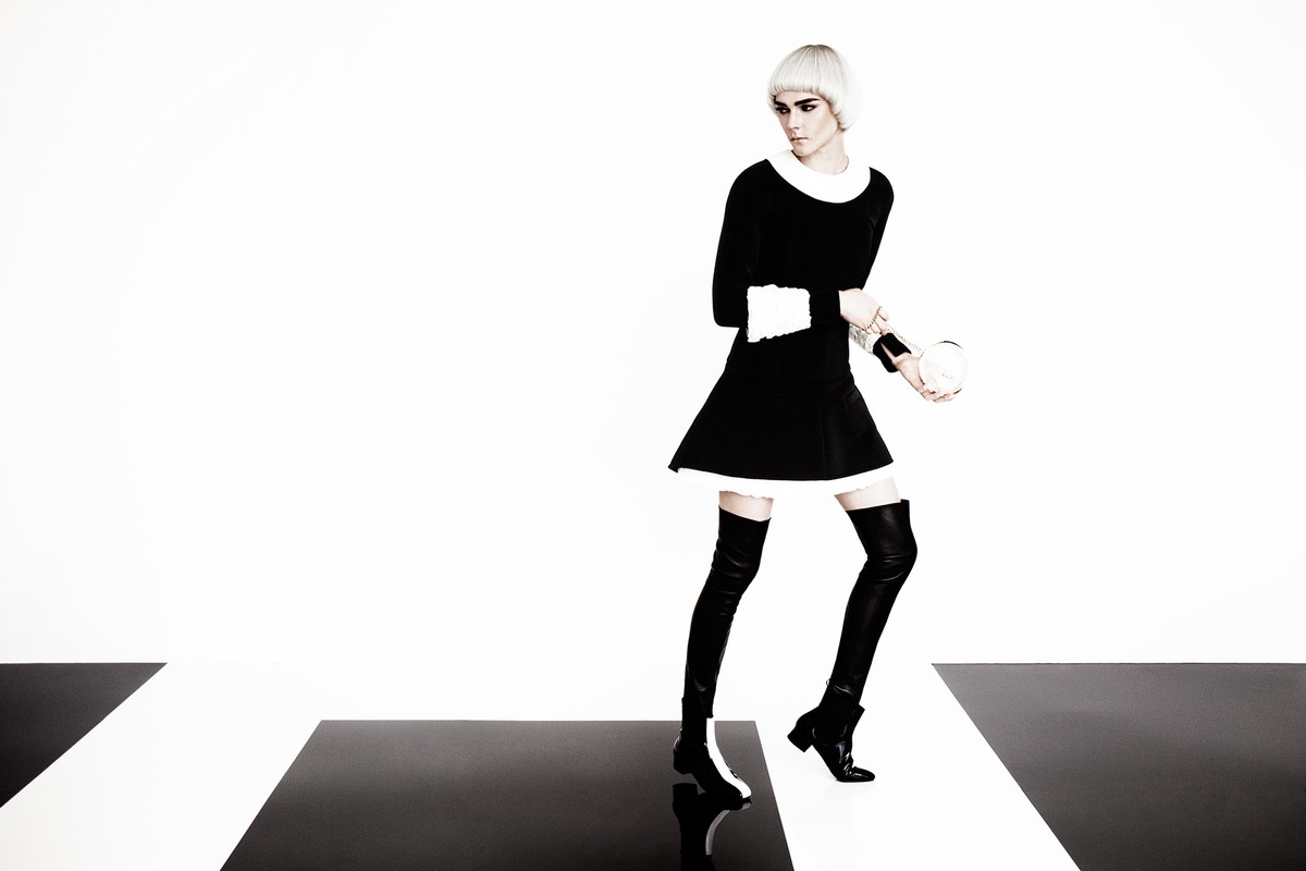 A slender adult fashion model poses in a black dress, black boots, and platinum blonde wig against a stark black and white studio backdrop.