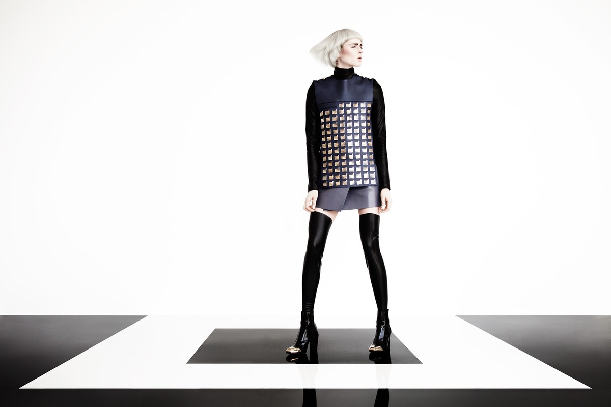 Fashion model poses in a studio wearing a navy and silver vest, gray skirt, black top, black thigh-high boots, and a short white haircut.
