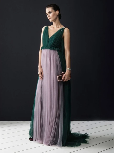 Elegant woman in a green and lilac tulle gown holding a clutch, posing against a dark background in a fashion-style portrait.