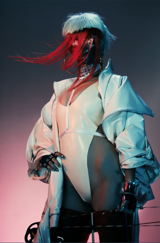 Stylish woman with futuristic mask, white jacket, and red hair, posing in a studio with blue and red background lighting.