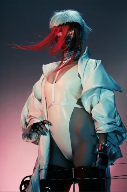 Stylish woman with futuristic mask, white jacket, and red hair, posing in a studio with blue and red background lighting.