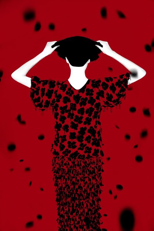 Back view of a person in a red and black floral dress with fringe, raising a black hat in a studio against a red background.