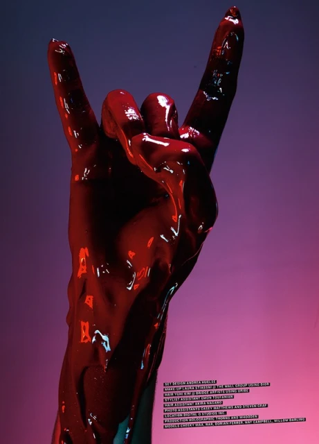 Hand with red paint in the shape of devil horns against a pink and blue background, indoor shot, artful manipulation of color.