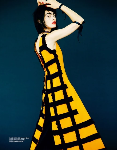 Fashion editorial shot of a female model with dark hair wearing a yellow sleeveless dress with black square lattice pattern, indoor against dark blue background.