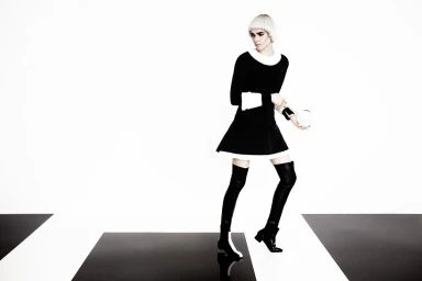 Model with white bob haircut wearing black dress with white trim and thigh-high boots against white background