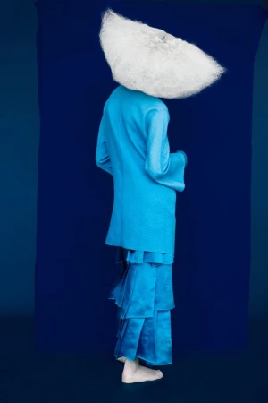Model seen from behind wearing a blue jacket and ruffled skirt with a massive white textured hat against a deep blue backdrop