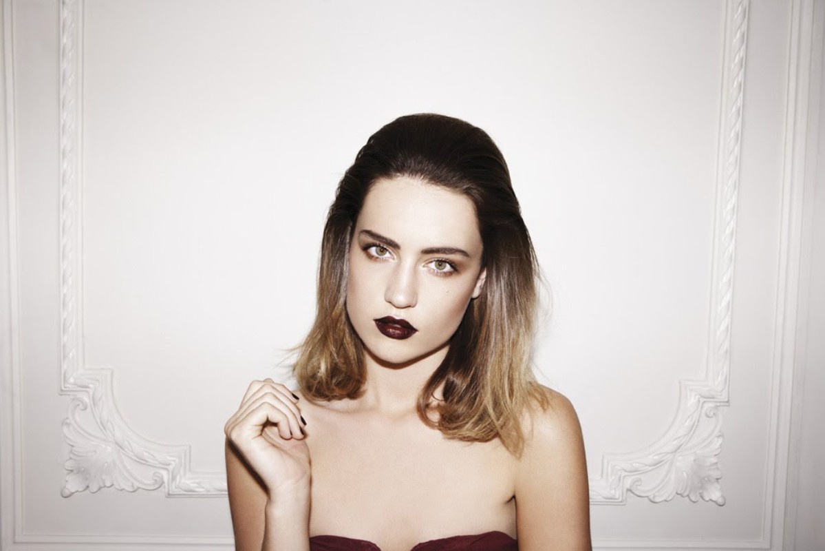 Portrait of a young woman with dark lipstick and shoulder-length hair against a white wall with ornamental molding.