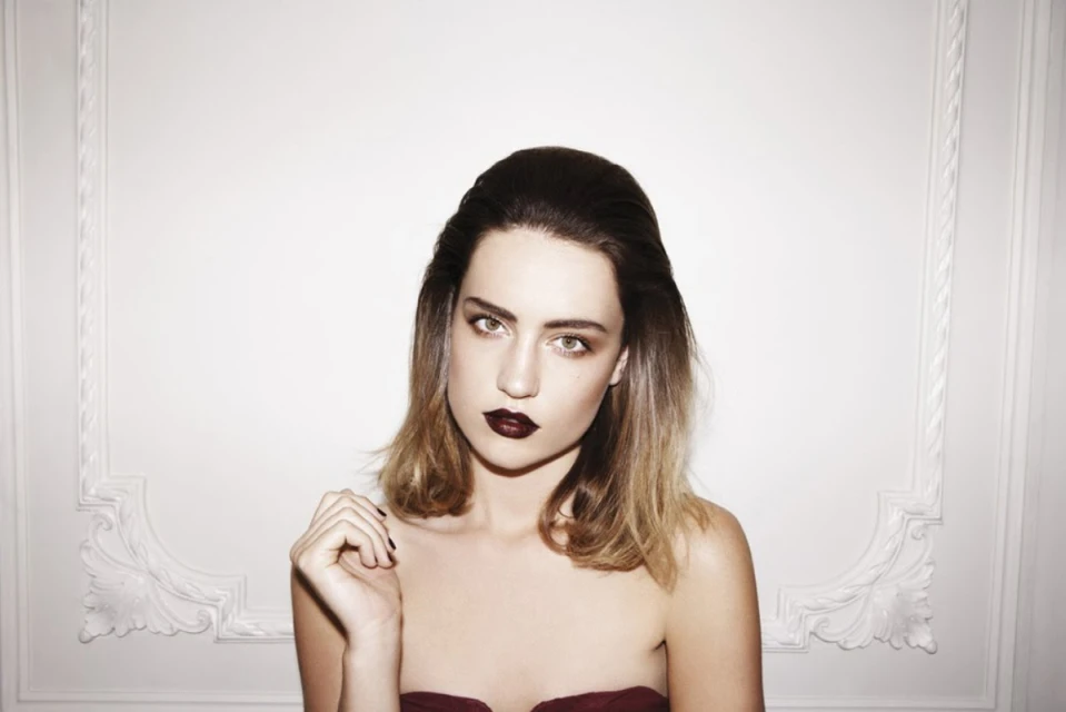 Portrait of a young woman with dark lipstick and shoulder-length hair against a white wall with ornamental molding.