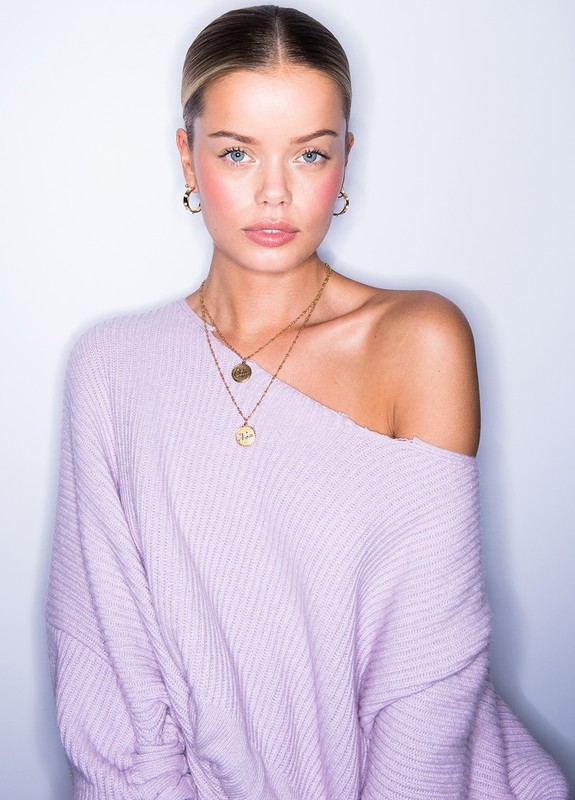 Portrait of a blonde woman in a purple sweater wearing gold jewelry, set against a clean white backdrop in a photo studio.