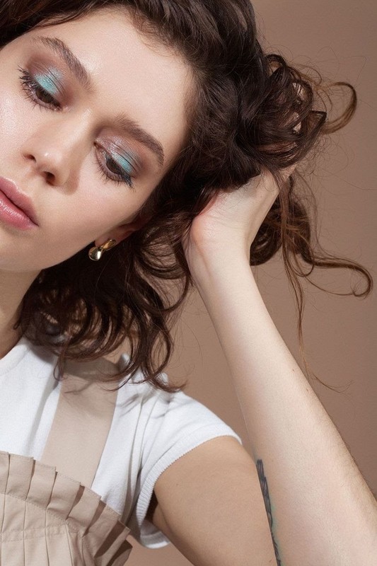 Close-up of an adult woman with brunette curly hair and light blue eyeshadow posing in a studio with a neutral background.
