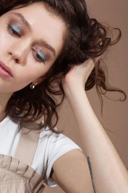 Close-up of an adult woman with brunette curly hair and light blue eyeshadow posing in a studio with a neutral background.