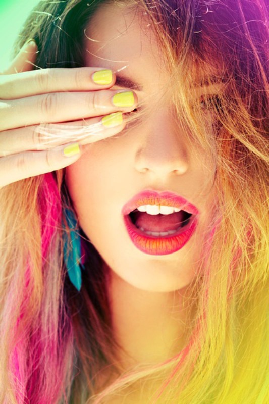 Close-up shot of a woman with vibrant hair and yellow nails covering her eyes, mouth slightly open showing teeth. Bright, colorful portrait.