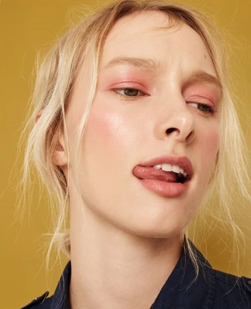 Close-up portrait of a blonde woman with pink eyeshadow sticking out her tongue against a yellow background