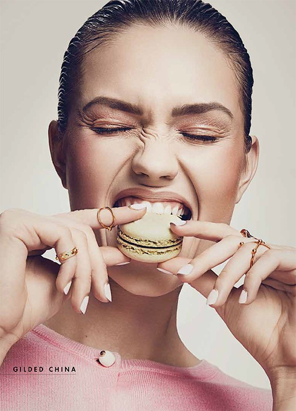 A young woman with slicked-back hair bites into a cream and brown macaroon, wearing rings and a pink sweater indoors in a studio.