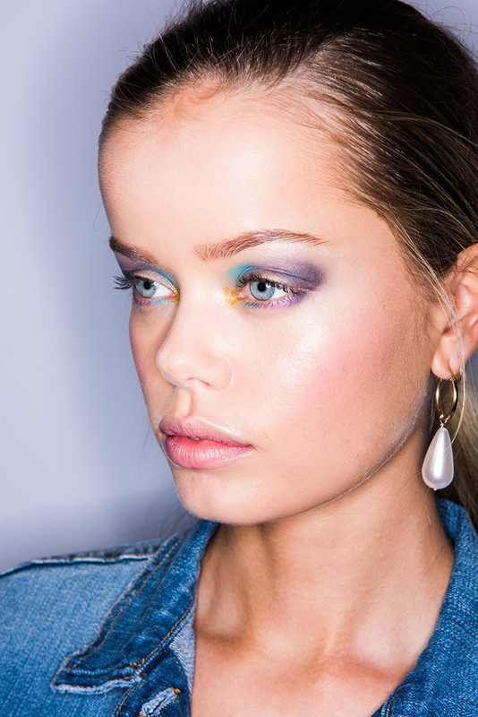 Close-up portrait of a young woman with colorful eye makeup, hoop earrings, and a denim jacket against a gradient grey background.