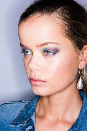 Close-up portrait of a young woman with colorful eye makeup, hoop earrings, and a denim jacket against a gradient grey background.