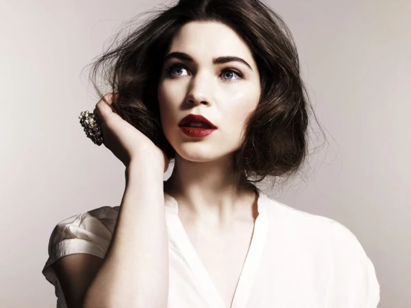 Close-up portrait of a woman with dark wavy hair, red lipstick, and a large beaded ring, wearing a white top against a neutral background.
