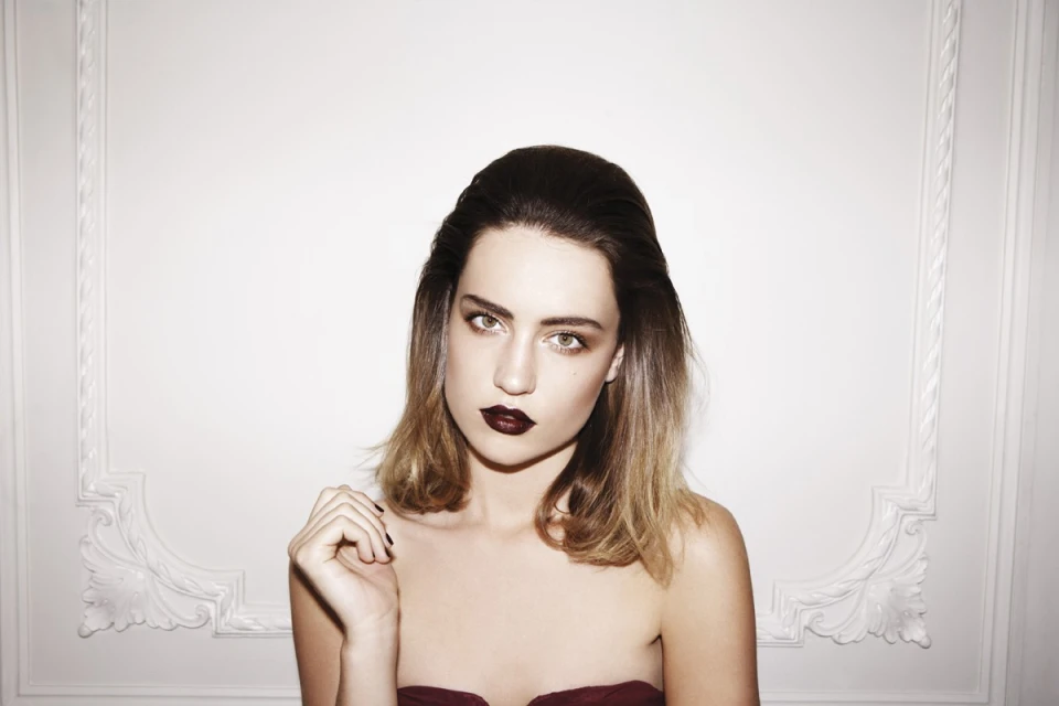 Portrait of a woman with dark lipstick and shoulder-length brown hair, against a white wall with ornate molding.