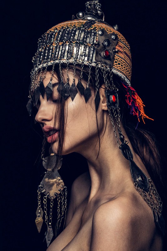 Adult female poses in a profile shot wearing an ornate, vintage headdress with metallic jewelry details against a dark background indoors.