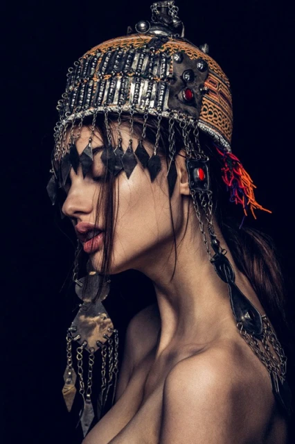 Adult female poses in a profile shot wearing an ornate, vintage headdress with metallic jewelry details against a dark background indoors.