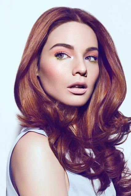 Portrait of an adult female with auburn wavy hair, wearing makeup, posing in a light blue top against a light backdrop in a photo studio.