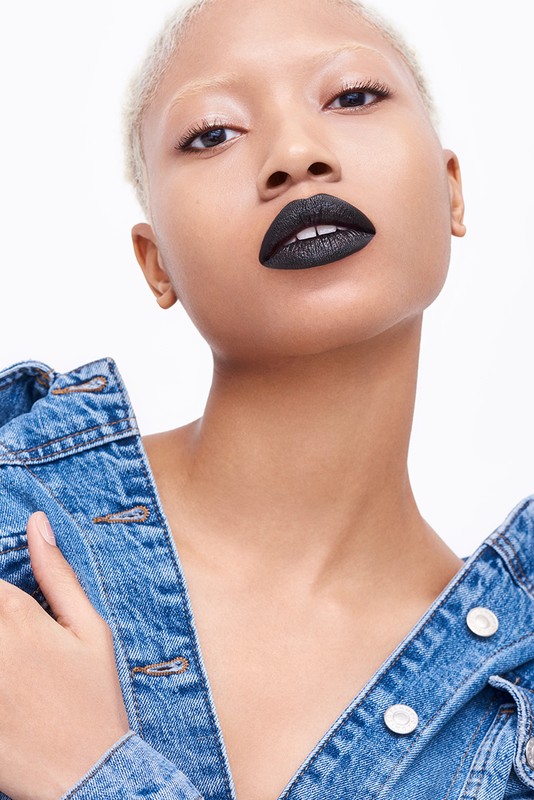 Close-up portrait of a young adult woman with platinum blonde hair, wearing a denim jacket and black lipstick against a white background.
