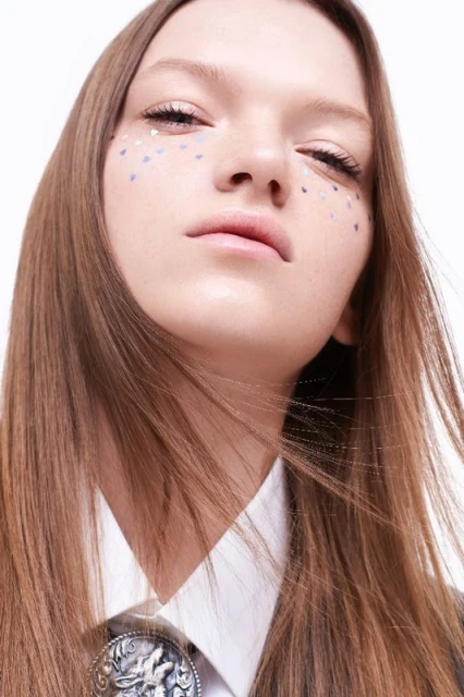 Close-up shot of a young woman with light brown hair and light skin, adorned with silver heart-shaped glitter freckles.