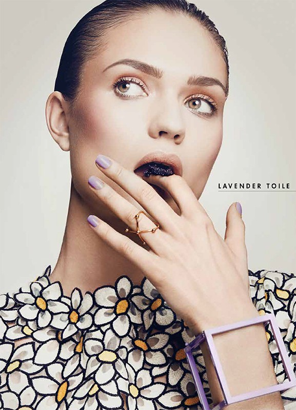 Eye-level portrait of a woman with lilac nails, a patterned dress, and makeup, holding a finger up to her dark-stained lips.