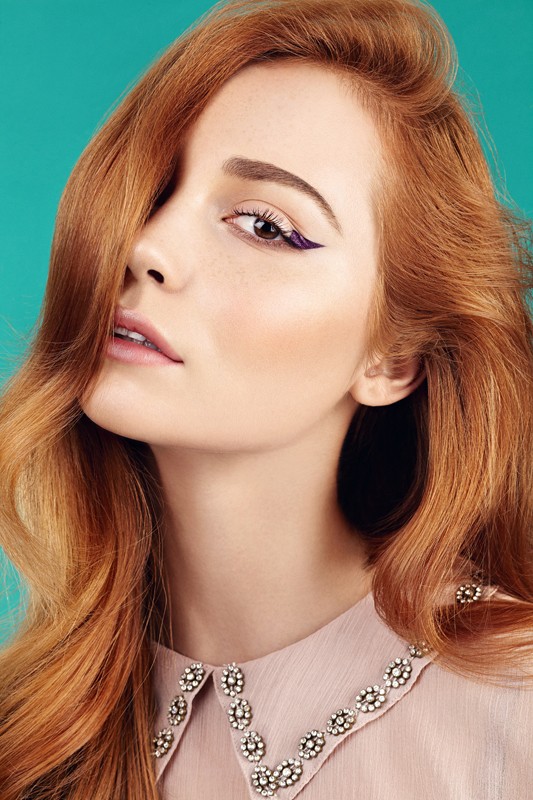 Close-up studio shot of a redhead woman with flawless skin, wearing a blush-colored blouse with a beaded collar, against an aqua background.