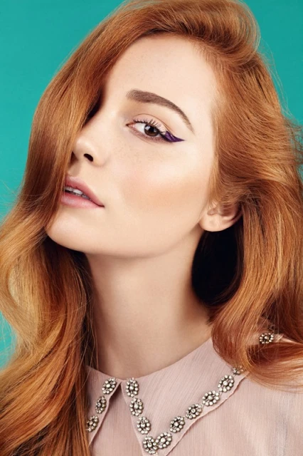 Close-up studio shot of a redhead woman with flawless skin, wearing a blush-colored blouse with a beaded collar, against an aqua background.