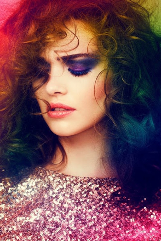 Close-up portrait of a woman with curly hair and dramatic purple eye makeup, wearing a sequined top with a colorful light effect.
